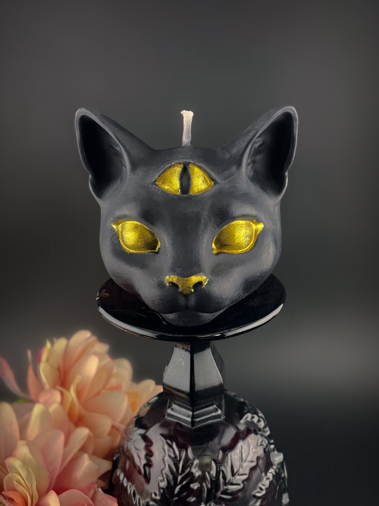 A black cat-shaped candle with metallic gold eyes, nose, and forehead mark, displayed on a black stand against a dark gray background with a soft peach flower in the lower left corner.