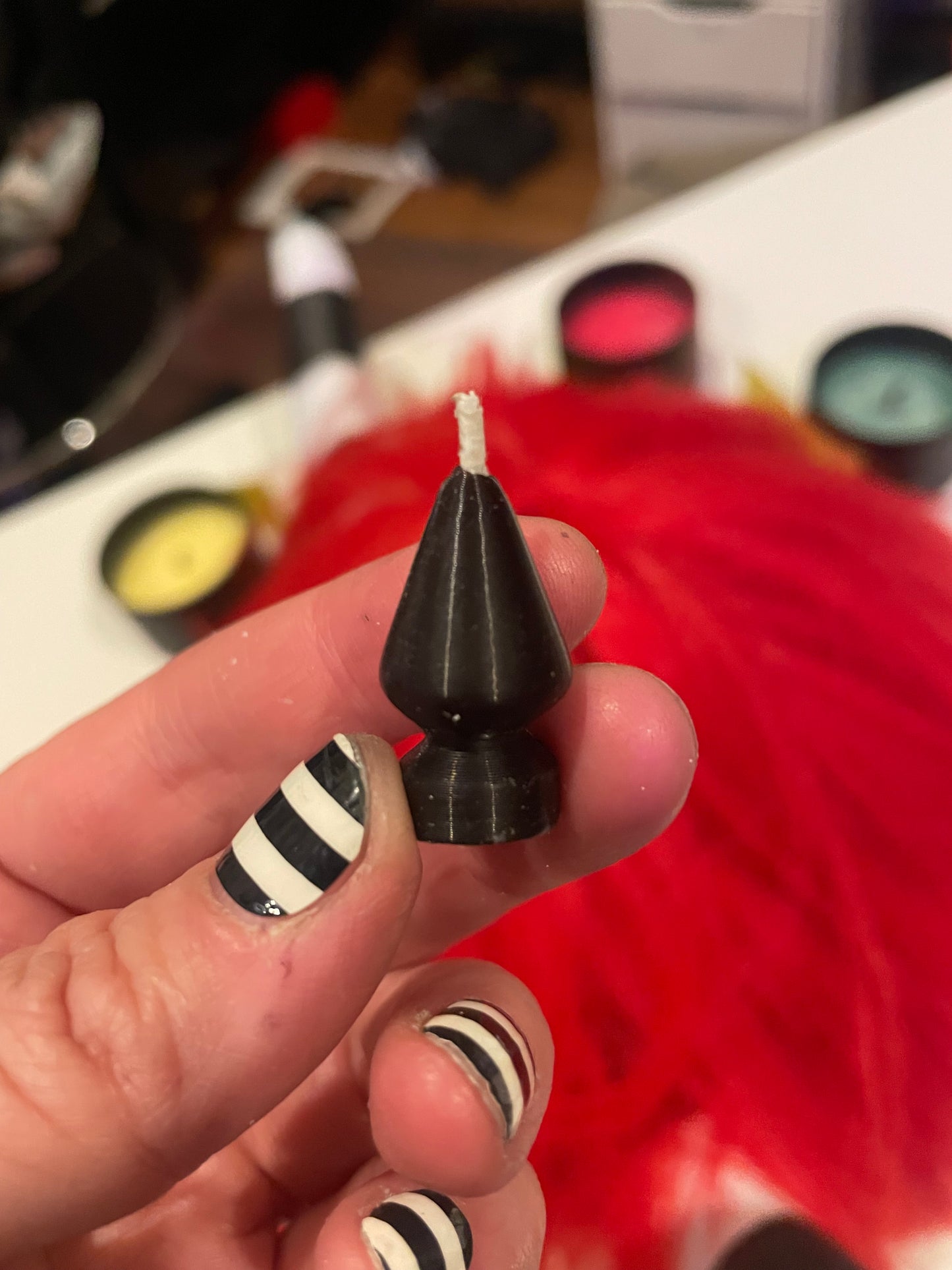 A hand with black and white striped nail art holds a small black cone-shaped butt plug candle with a white wick, with a bright red decoration in the blurred background.