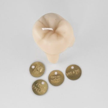 A beige novelty candle shaped like a clenched fist, shown with four round brass tokens with text that reads "FIGHT FASCISM", "BREATHE", "DRINK WATER", "EAT THE RICH" on a plain light gray background.