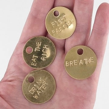 Four round brass tokens with hand-stamped phrases are displayed on a person's palm. The phrases read 'FIGHT FASCISM', 'EAT THE RICH', 'DRINK WATER', and 'BREATHE'.