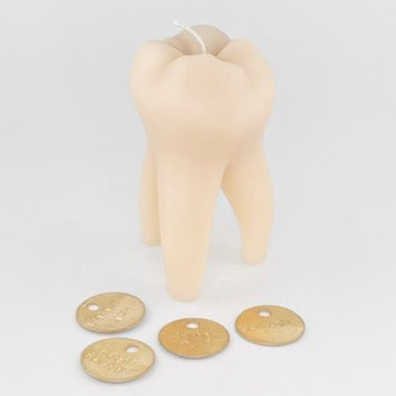 A novelty tooth-shaped candle in a light beige color, shown with four small brass tokens of advice on a plain light gray background.