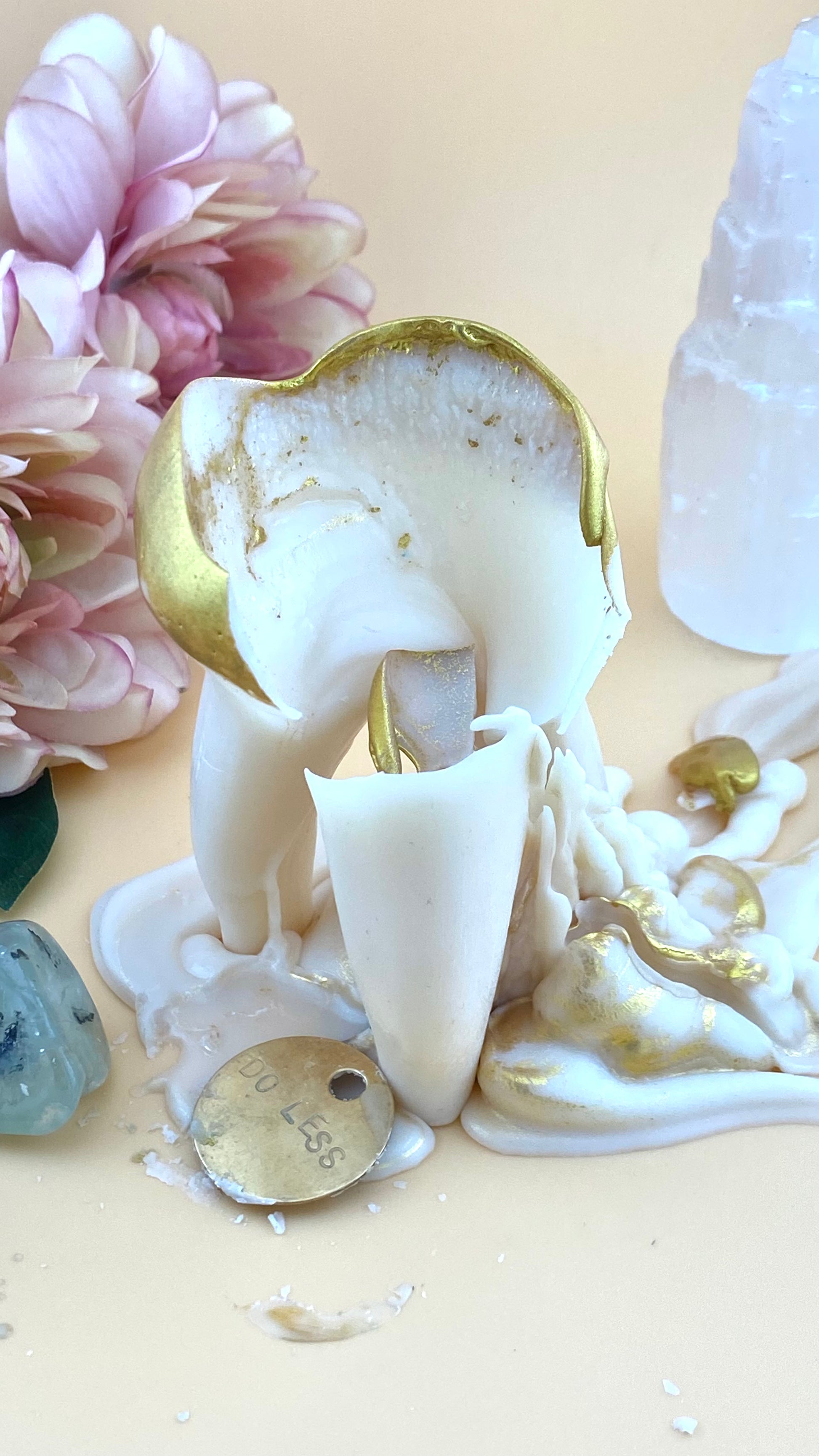 A melted tooth candle with gold embellishments and dripping wax, styled with pink flowers, crystals, and a small brass token that reads "DO LESS" on a beige background.