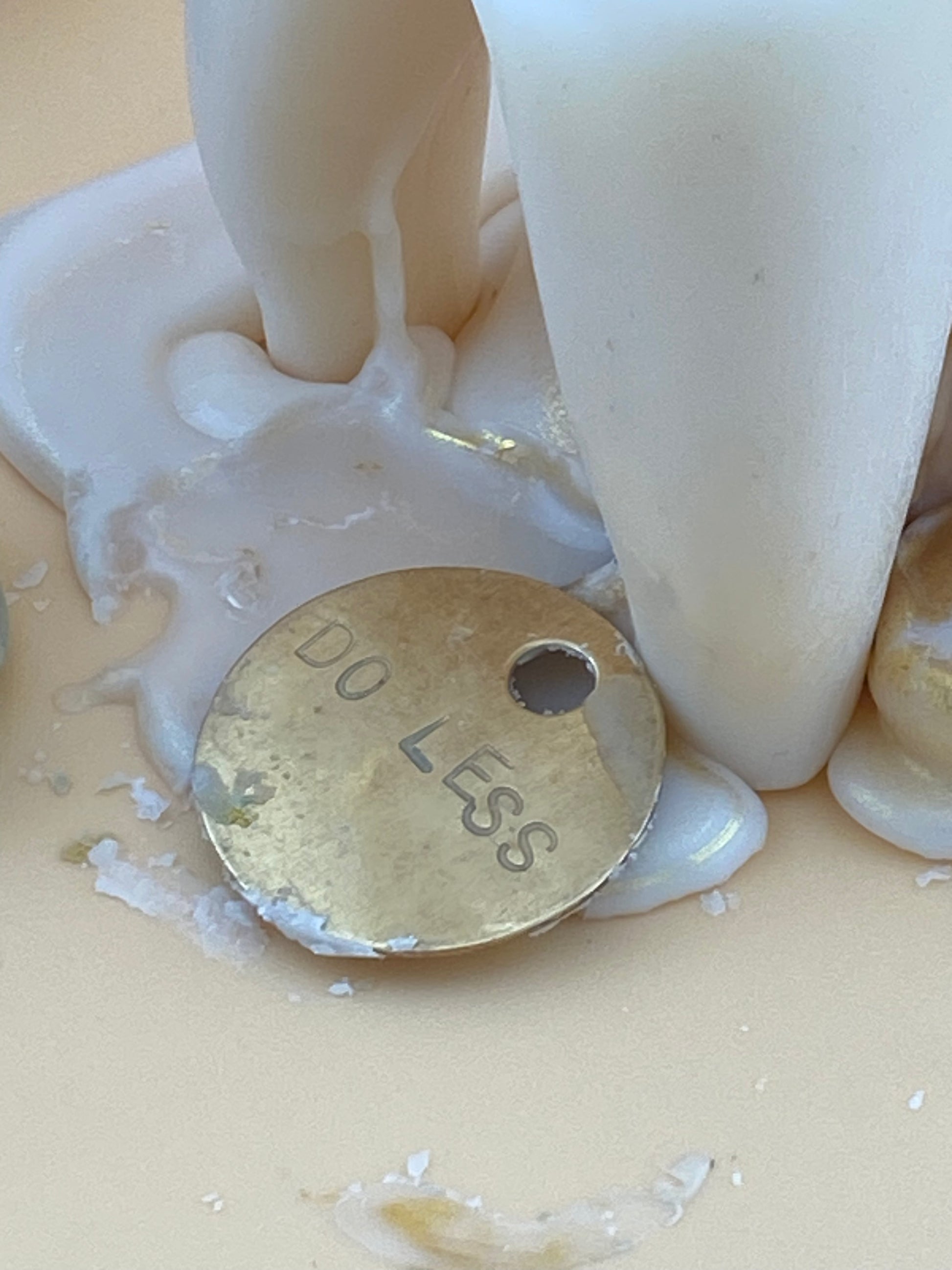 A round brass token hand-stamped with 'DO LESS', resting on a beige surface surrounded by melted white wax.