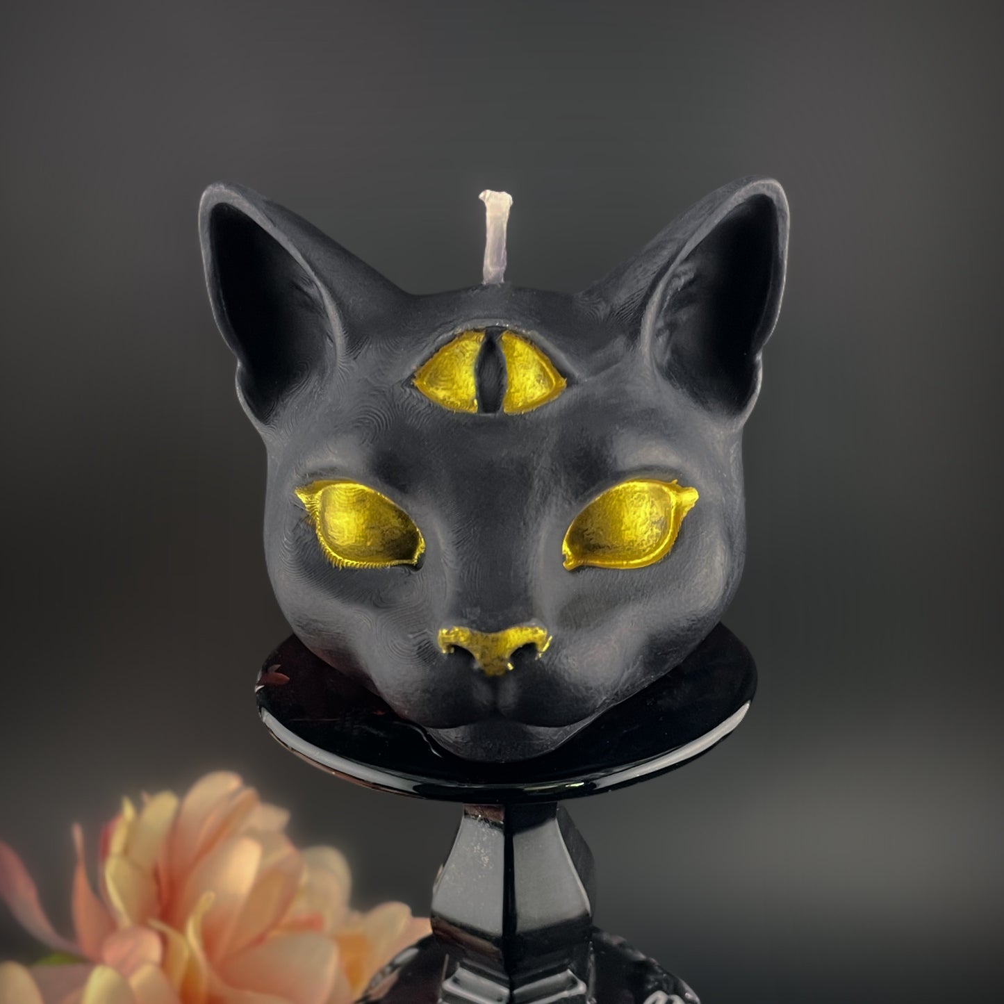 A black cat-shaped candle with metallic gold eyes, nose, and forehead mark, displayed on a black stand against a dark gray background with a soft peach flower in the lower left corner.