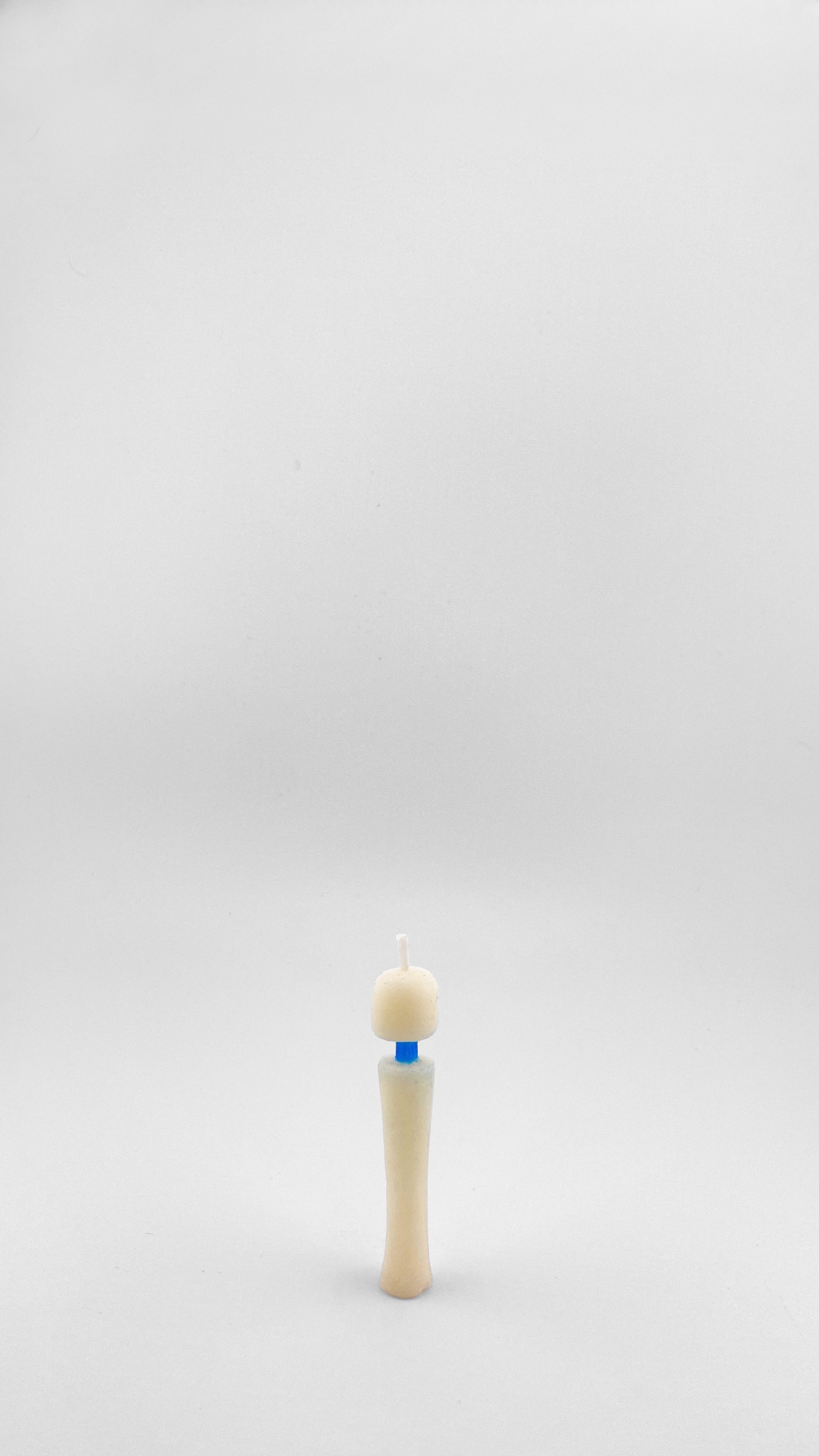A small novelty candle, the size of a birthday candle, shaped like the iconic Magic Wand vibrator, made with off-white soy wax and small blue accent at the neck, set against a seamless light gray background.