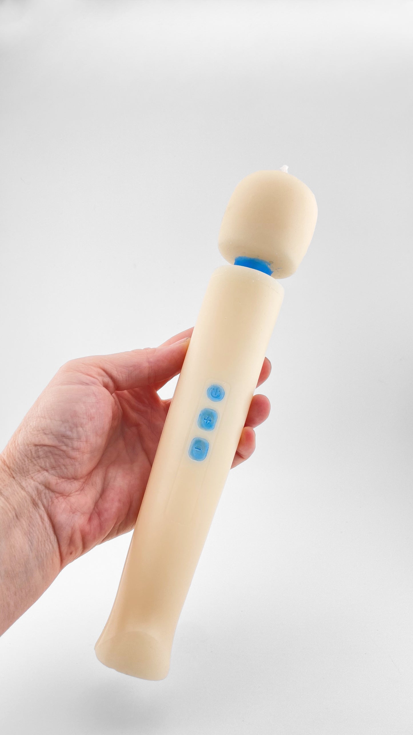 A hand holding an off-white candle that resembles the iconic Magic Wand  personal massager vibrator with blue buttons and a blue neck, set against a light gray background.