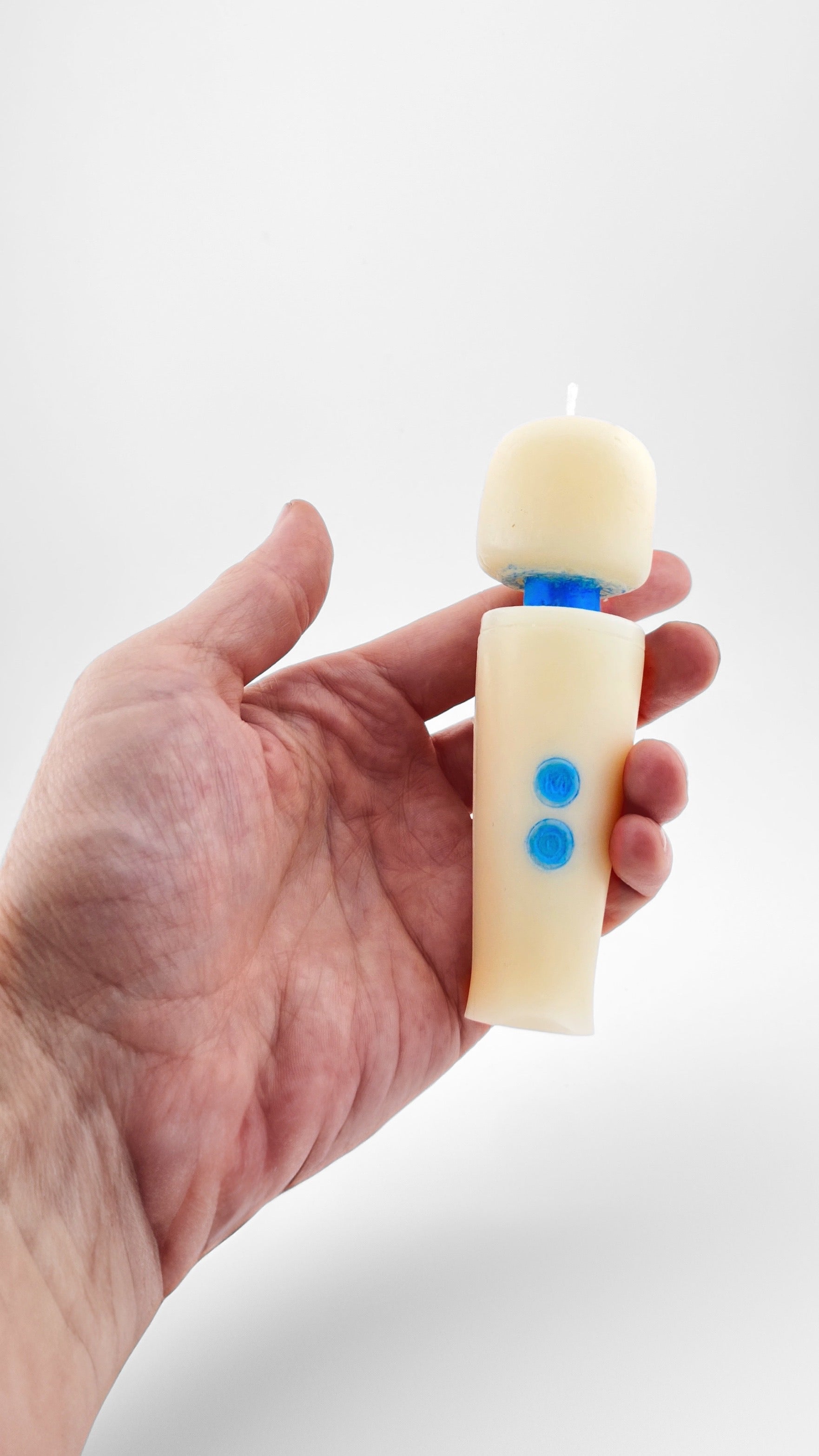 A hand holding a novelty candle shaped like a miniature Magic Wand, made with off-white soy wax with a blue neck and buttons, against a plain light gray background.