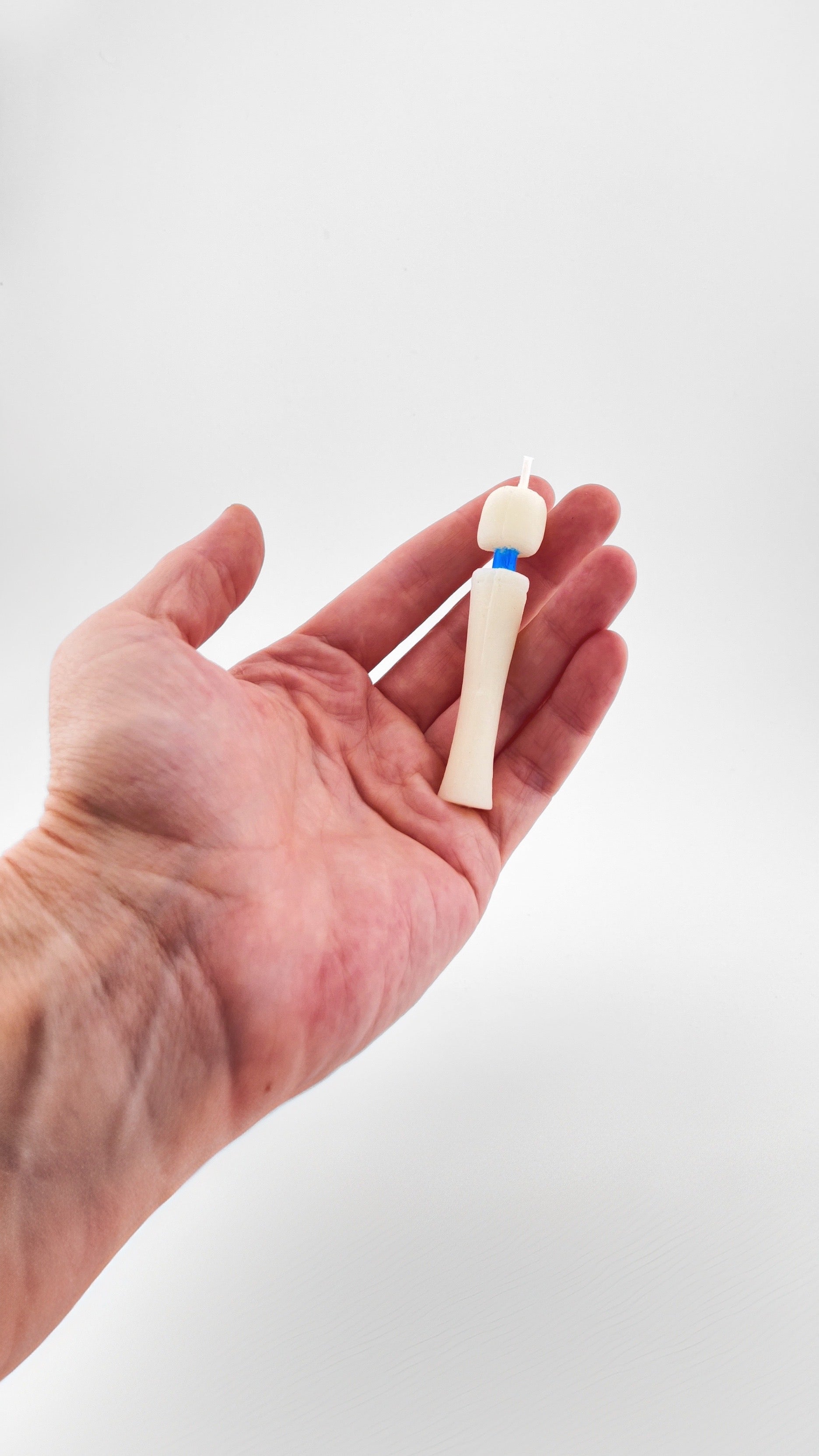 A hand holding a birthday candle sized novelty candle shaped tiny Magic Wand vibrator made with off-white soy wax with a blue neck and white wick, set against a plain light gray background.
