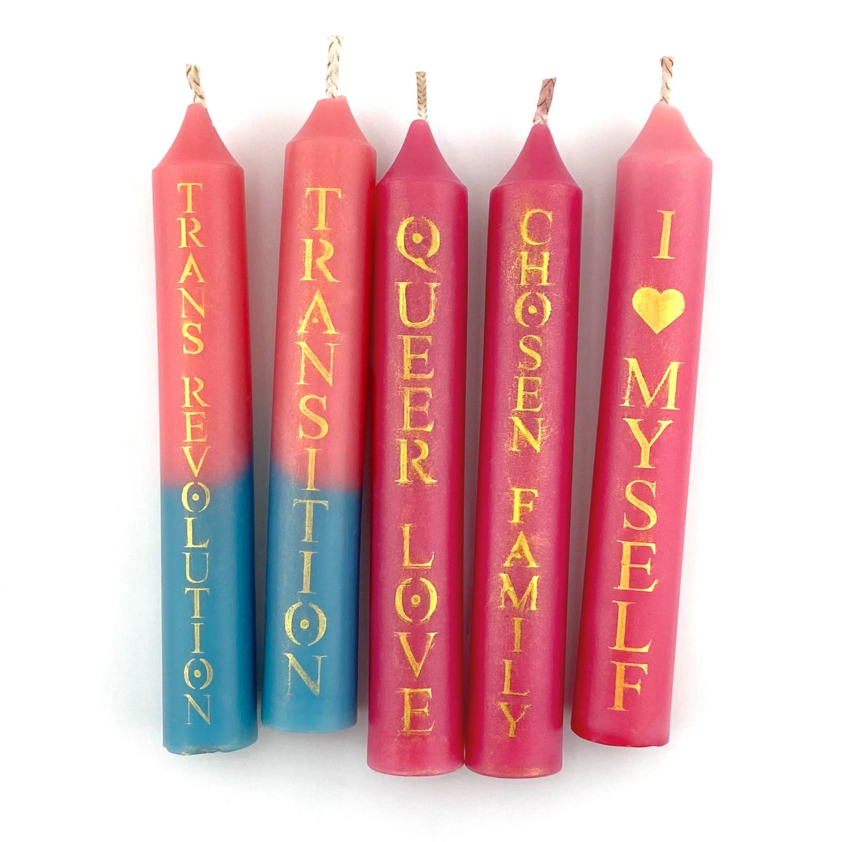 Five taper candles on a white background, each with printed phrases in gold lettering. Three are pink with the text reading'QUEER LOVE,' 'CHOSEN FAMILY,' and 'I ♥ MYSELF.' The other two candles have a pink-to-blue gradient and read 'TRANSITION' and 'TRANS REVOLUTION'.