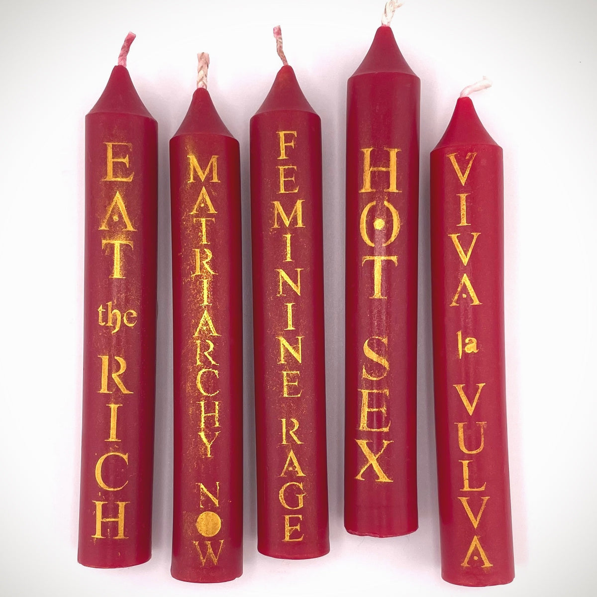 Five red taper candles with gold vertical text, displayed against a plain white background. The text reads 'EAT the RICH', 'MATRIARCHY NOW', 'FEMININE RAGE', 'HOT SEX', and 'VIVA LA VULVA'.