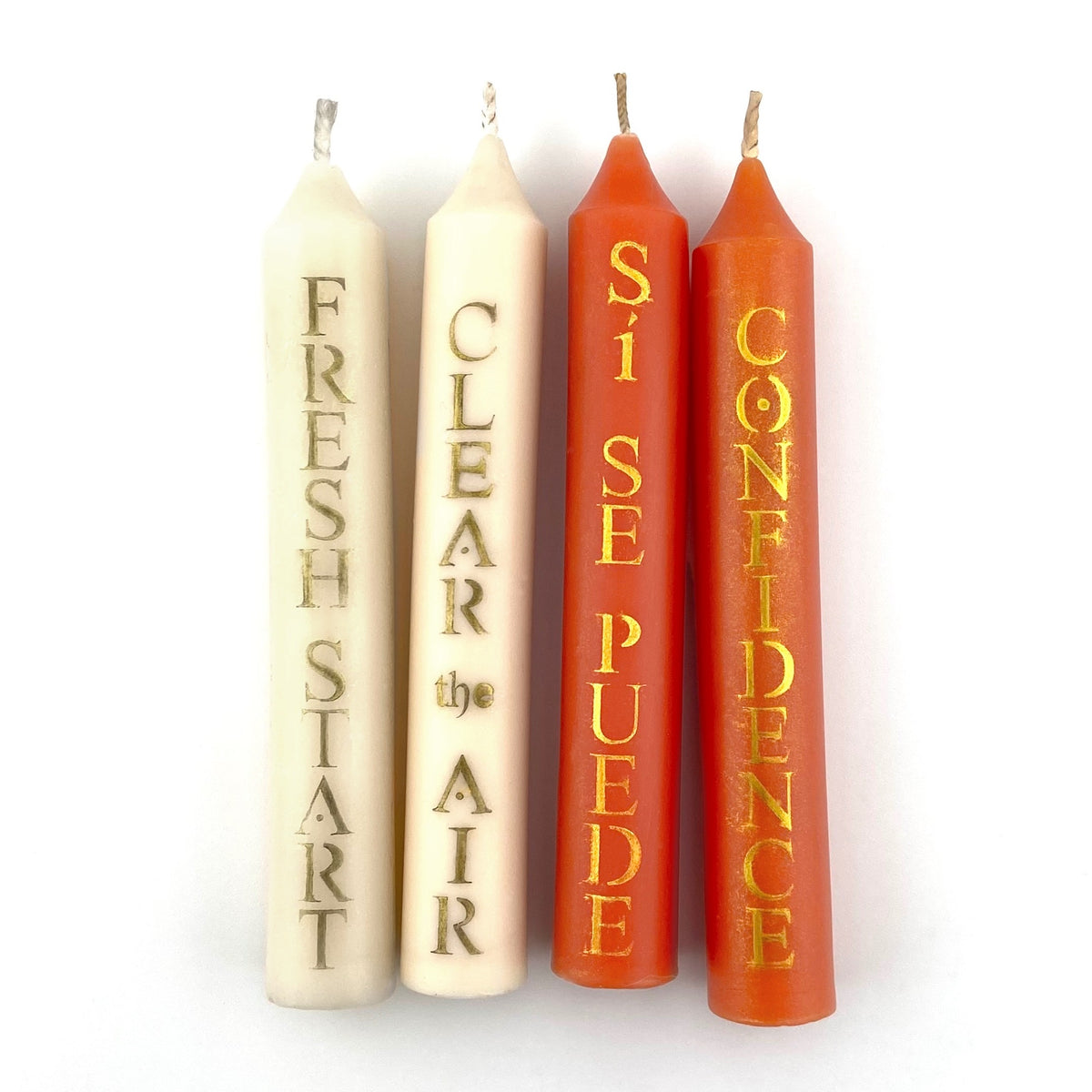 Four taper candles with text on a white background. Two candles are white with text reading FRESH START and CLEAR THE AIR. The other two candles are orange and read SI SE PUEDE and CONFIDENCE.