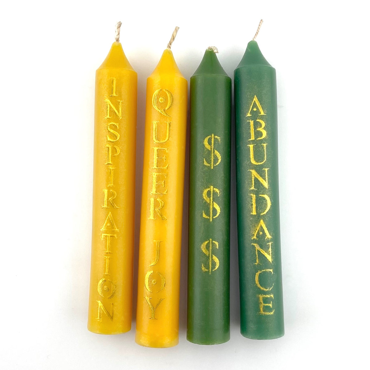 Four taper candles in yellow and green with gold text on a white background. The two yellow candles say INSPIRATION and QUEER JOY. One of the green candles says ABUNDANCE and the other has three $ symbols.